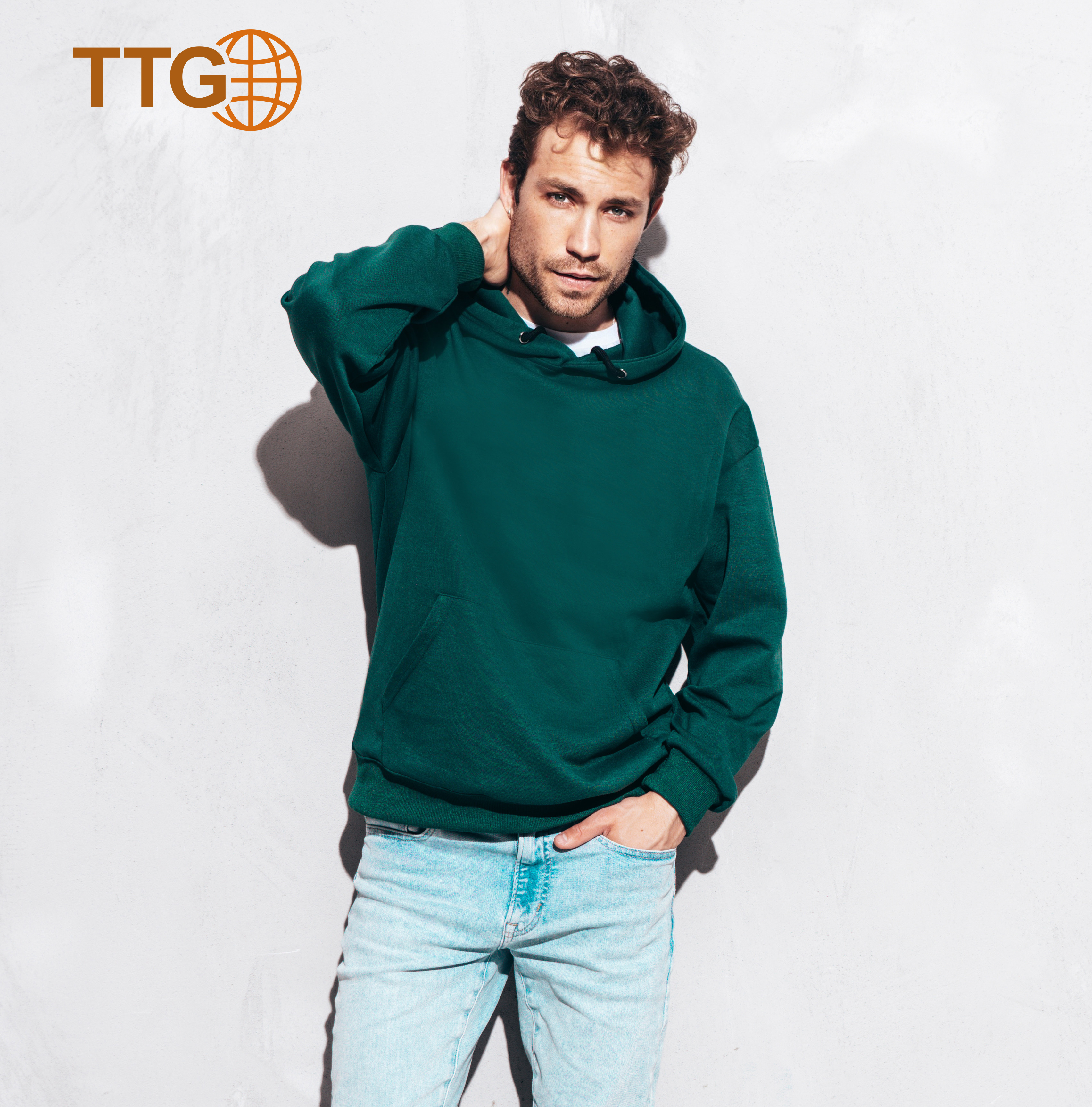 Handsome Confident Hipster  Model.sexy Unshaven Man Dressed In Summer Stylish Green Hoodie And Jeans Clothes. Fashion Male With Curly Hairstyle Posing In Studio. Isolated On Grey